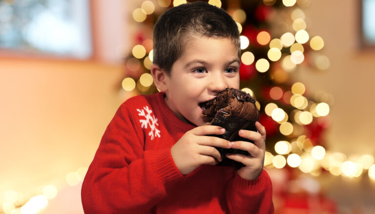 Un bambino felice che tiene un pandoro artigianale in mano, con uno sfondo natalizio illuminato da luci calde e un albero di Natale.